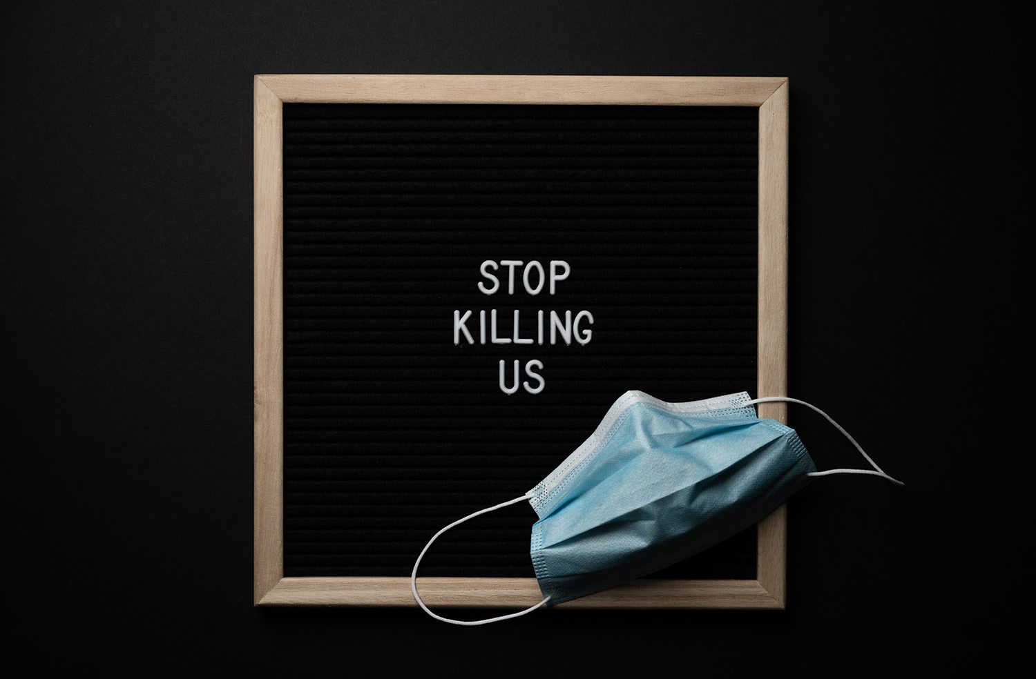 Photo of a disposable blue mask in front of a wooden sign with the words "Stop Killing Us"