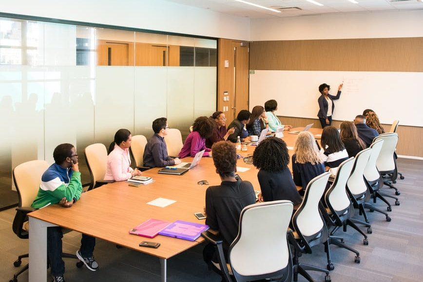 Picture of a boardroom table with lots of people around it and a person presenting on a whiteboard