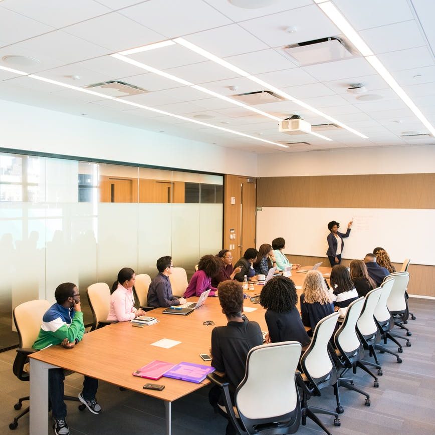 Picture of a boardroom table with lots of people around it and a person presenting on a whiteboard 