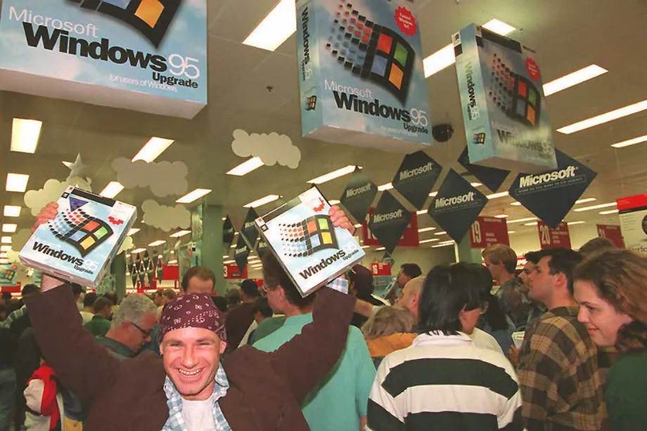 It is a photo of a sale scene. The room is full of people and there is a man in a brown jacket and a red bandanna holding up two Windows 95 boxes. There are big versions of Windows 95 boxes hanging from the ceiling