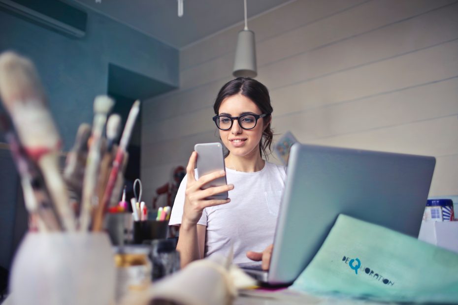 A largely decorative image of a woman with dark hair, she has glasses with thick black frames on. She is sitting at a messy desk in front of a laptop and she is smiling at her mobile phone.