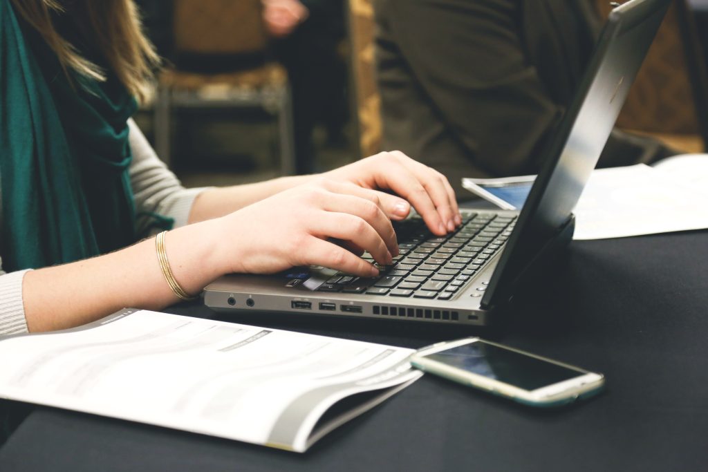 A largely decorative image of a woman wearing a green top typing on a laptop. You can see other people behind her also working on laptops. There is a smart phone in the foreground.
