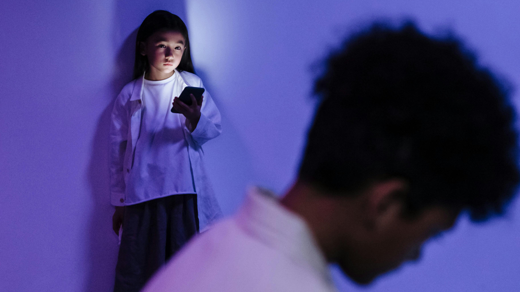 Child holding a phone, looking at an out-of-focus shadowy figure in the foreground. The light from the phone is lighting her up in a dark room infused with purple light. This is a decorative image intended to show that sometimes young people have devices and they don't always feel safe.
