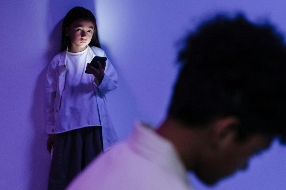 Child holding a phone, looking at an out-of-focus shadowy figure in the foreground. The light from the phone is lighting her up in a dark room infused with purple light. This is a decorative image intended to show that sometimes young people have devices and they don't always feel safe.