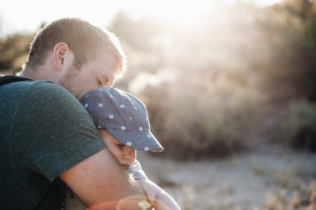 A largely decorative image of a man holding a small baby in a blue hat. The background is blurry, but it looks like they are outside.