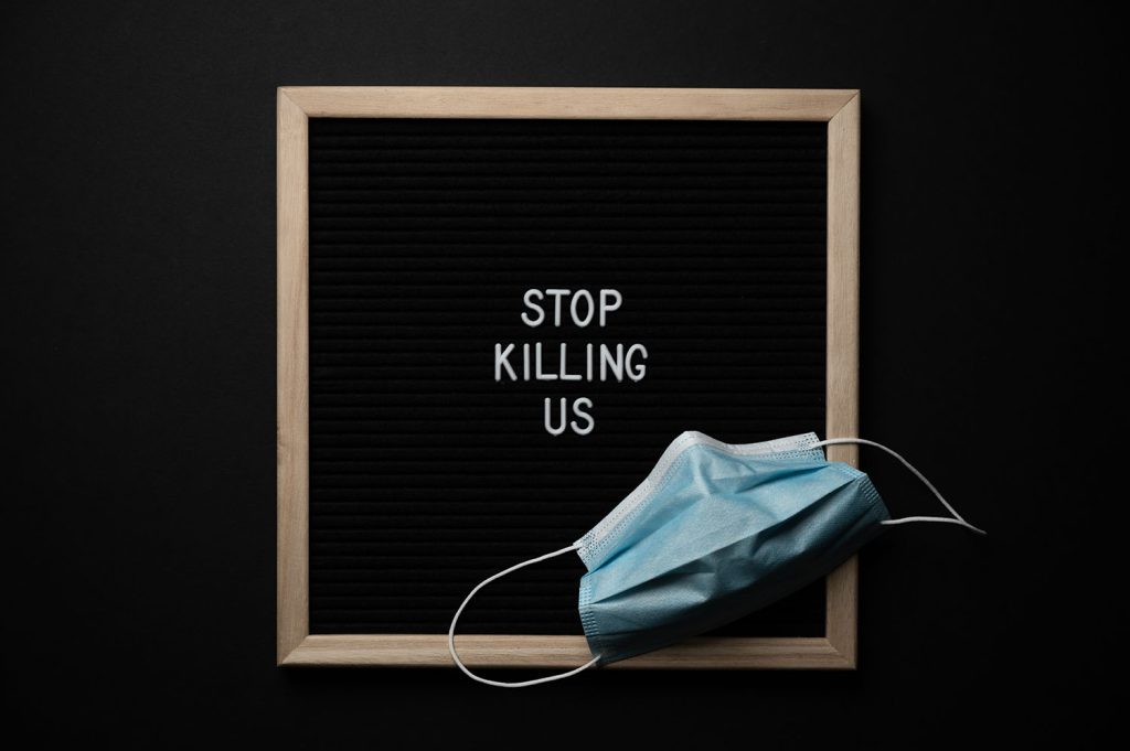 Photo of a blue medical mask sitting on a letter board, the kind that you push individual letters into a velvet holder. The words on the board say "Stop Killing Us". This is a largely decorative image meant to provoke strong feelings.