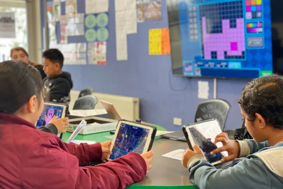 Photo of kids looking at tablets and there is some kind of colourful coding activity on the screen at the front of the classroom