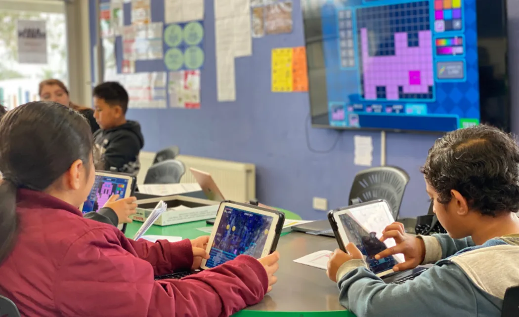 Photo of kids looking at tablets and there is some kind of colourful coding activity on the screen at the front of the classroom