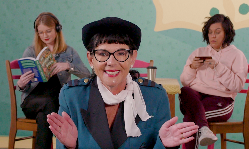 Photograph of comedian Michèle A'Court dressed as an air hostess, with Leonie Hayden and Alex Casey in the background looking board on their phones. The entire scene is of a cute colour kitchen table