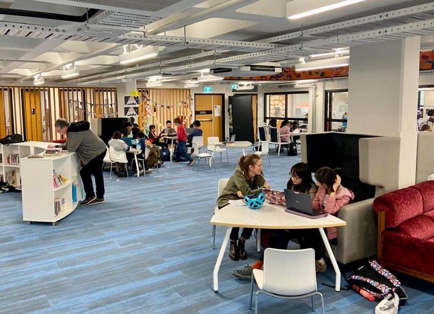 Photograph of a modern learning environment at a primary school. There are adults sharing large tables and the entire space is open