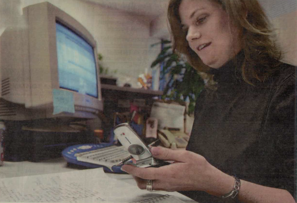 Photo of a woman using a desktop computer, holding a flip phone