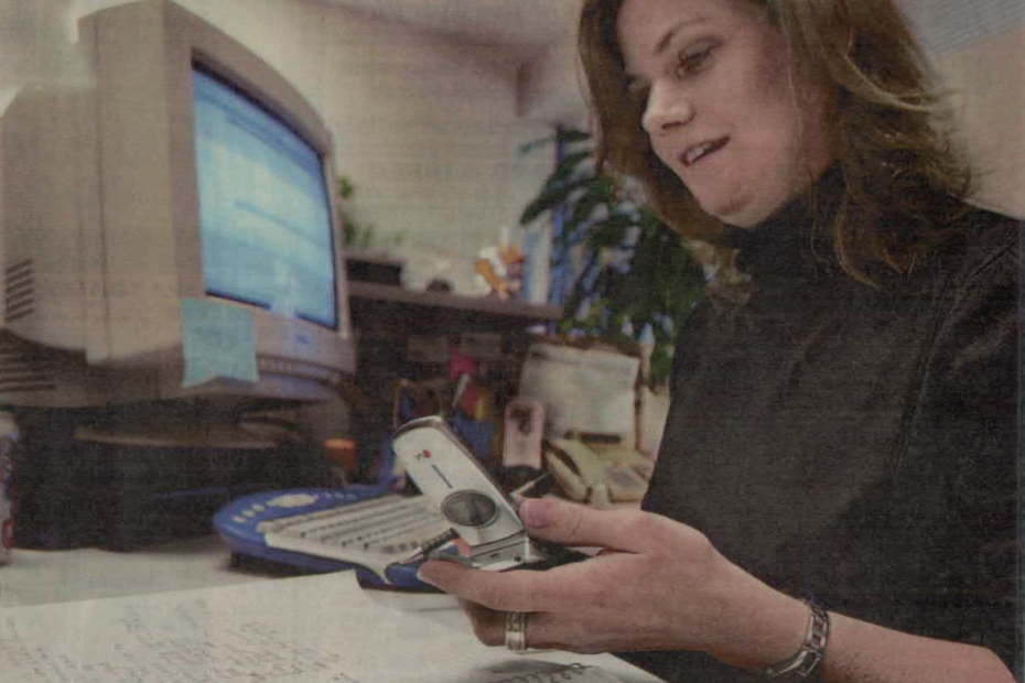 Photo of a woman using a desktop computer, holding a flip phone