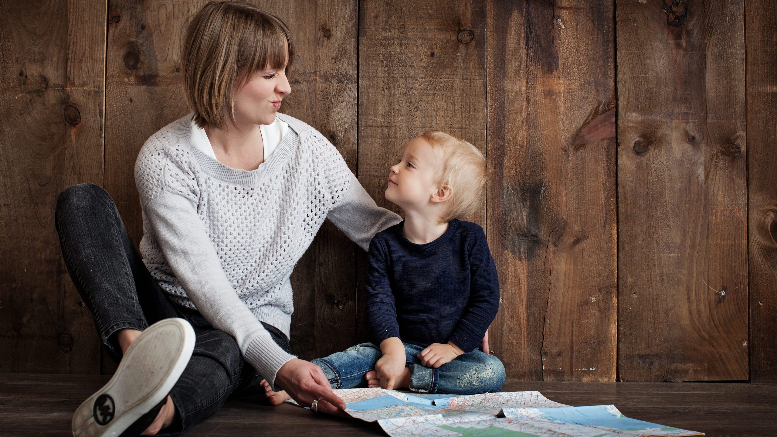 A largely decorative photograph, a stock photo of a woman making a cute face at a child. The two are sitting on the floor with a backdrop of a wooden wall, in front of a large paper map.